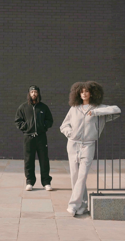 Two people wearing matching tracksuits standing against a dark brick wall.